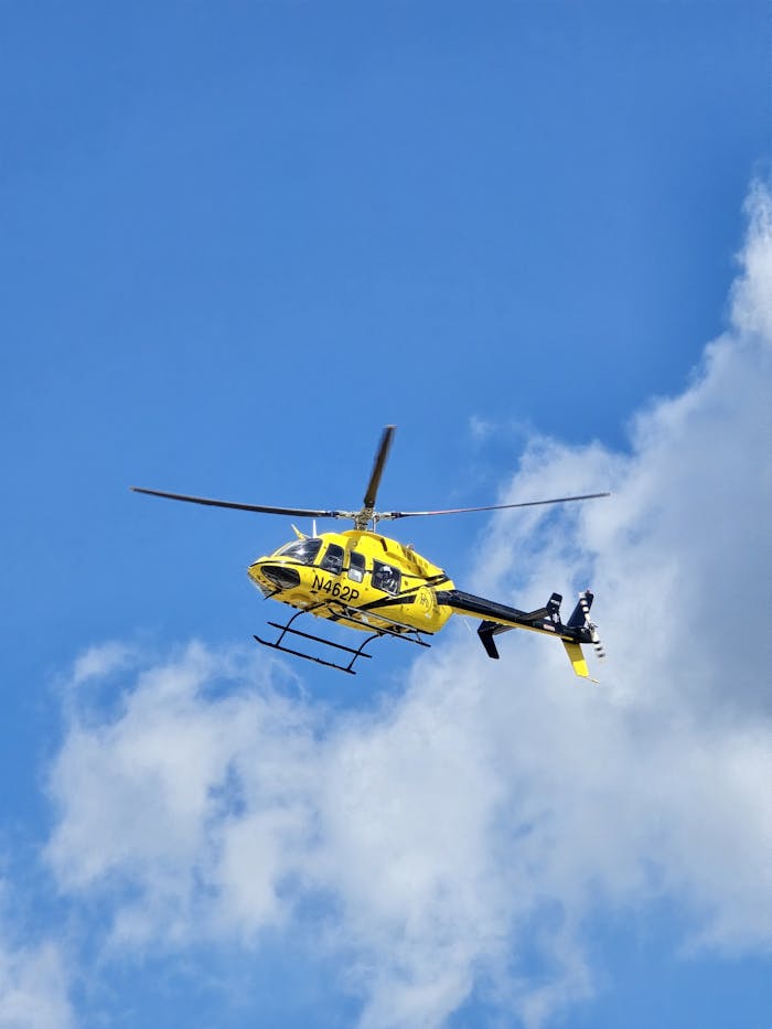 A yellow helicopter flying against a bright blue sky with scattered clouds.
